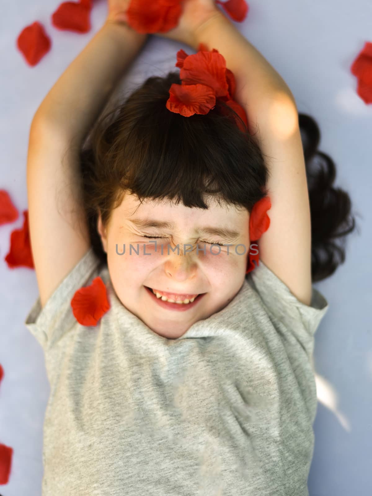 Smiling little girl squints as rose petals fall on her, vertical portrait