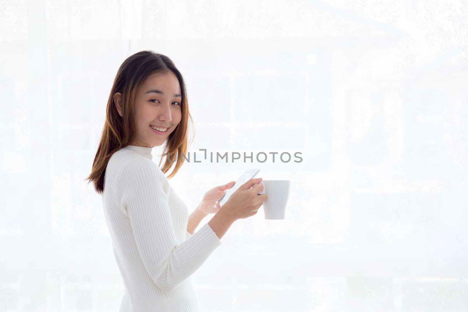 Beautiful young woman in white sweater drinking coffee and holding smartphone at a window.