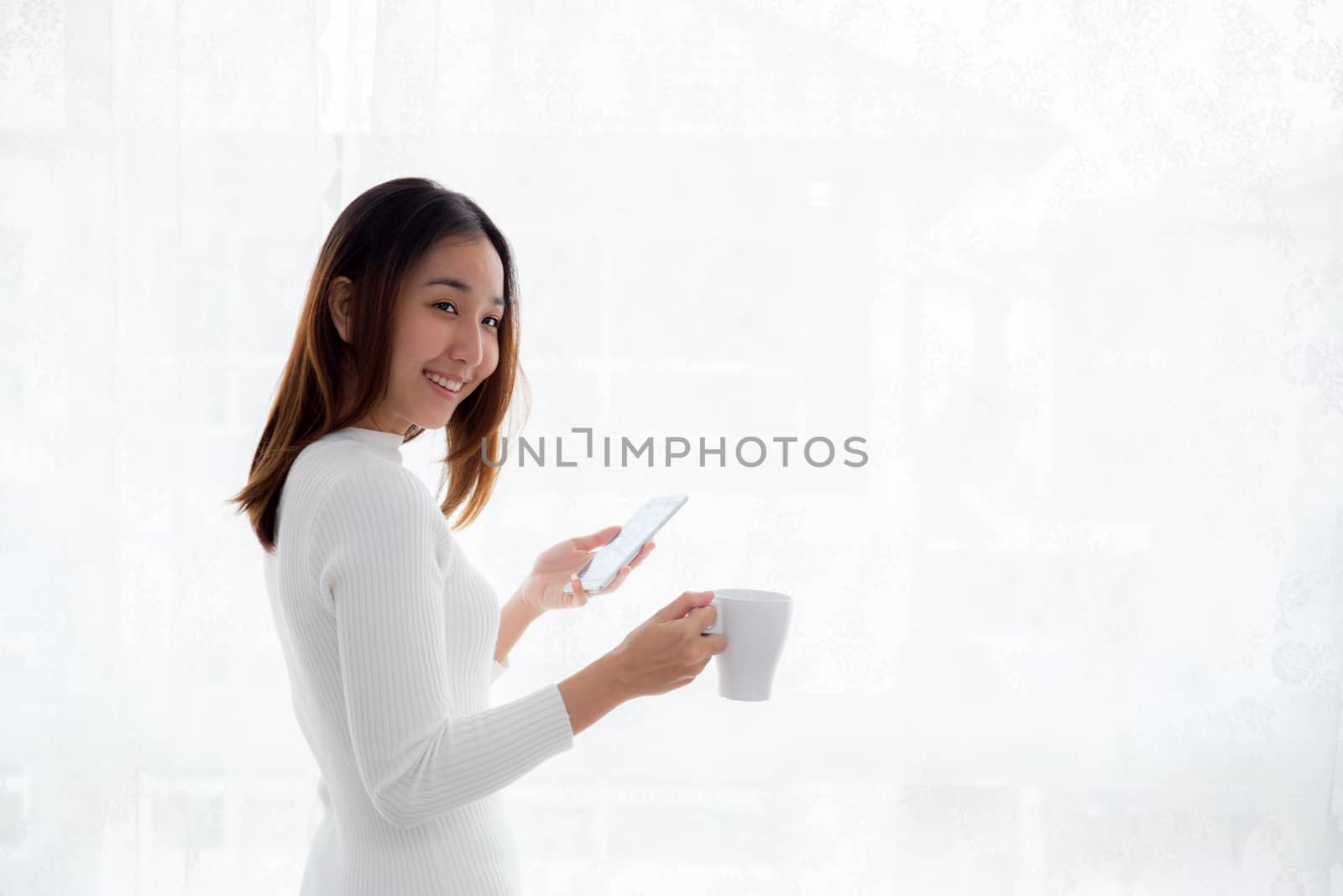 Beautiful young woman in white sweater drinking coffee and holding smartphone at a window.