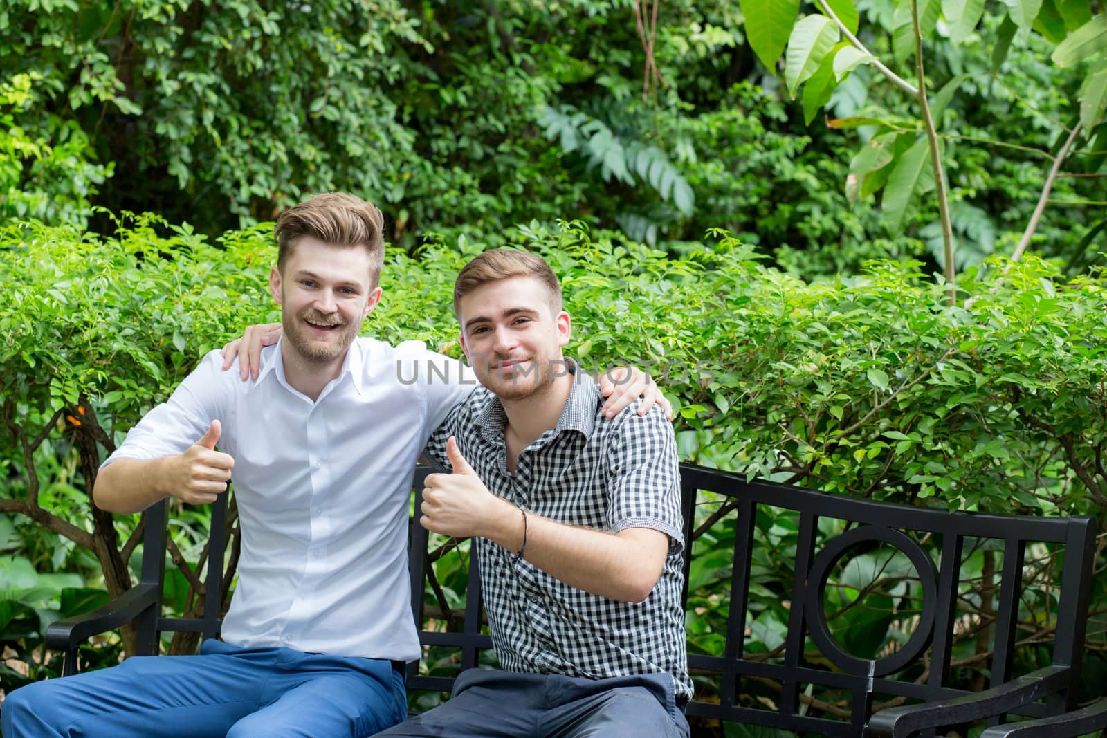 two happy young casual men making the ok thumbs up hand gesture in garden.