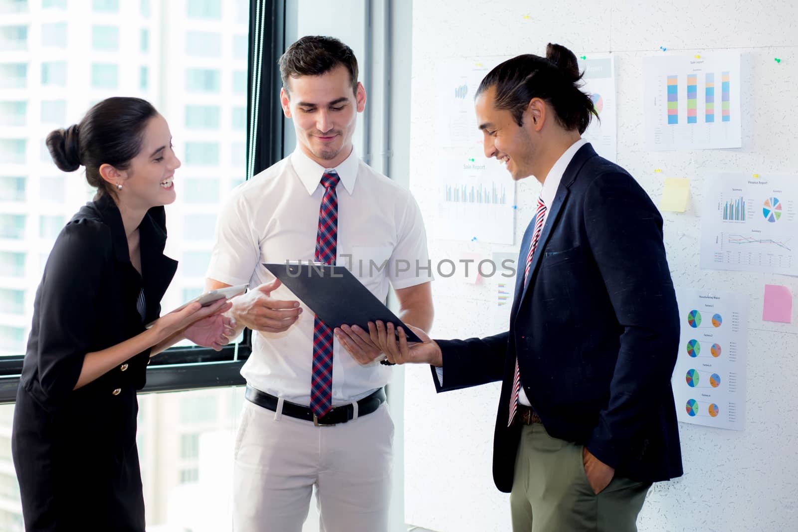 Three businesspeople standing in modern office looking file document and talking in meeting room.