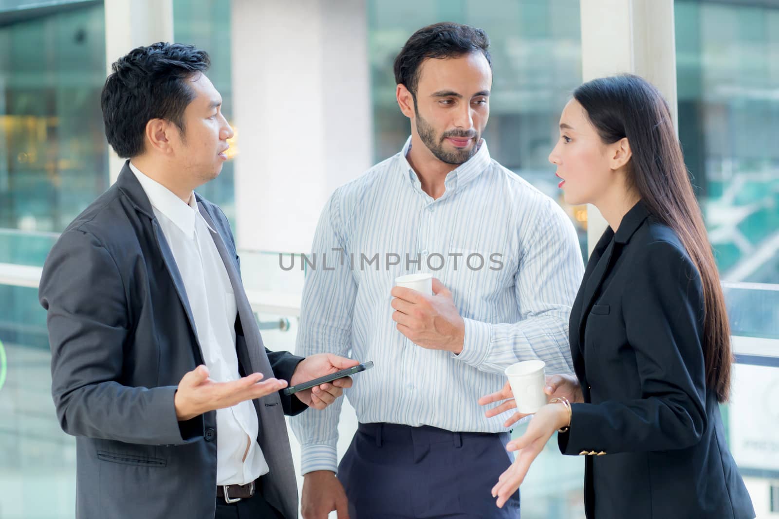 Two handsome young businessmen and lady in classic suits are holding cups of coffee, talking and smiling, standing outside the office building.