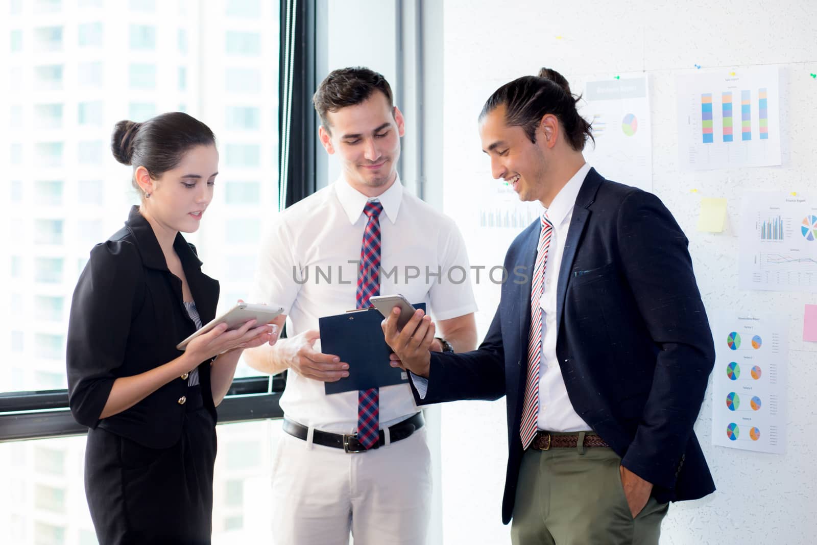 Three businesspeople standing in modern office looking at smart mobile phone and talking in meeting room.