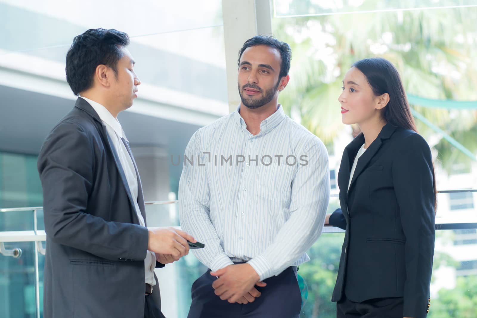 Two handsome young businessmen and lady in classic suits are holding cups of coffee, talking and smiling, standing outside the office building.