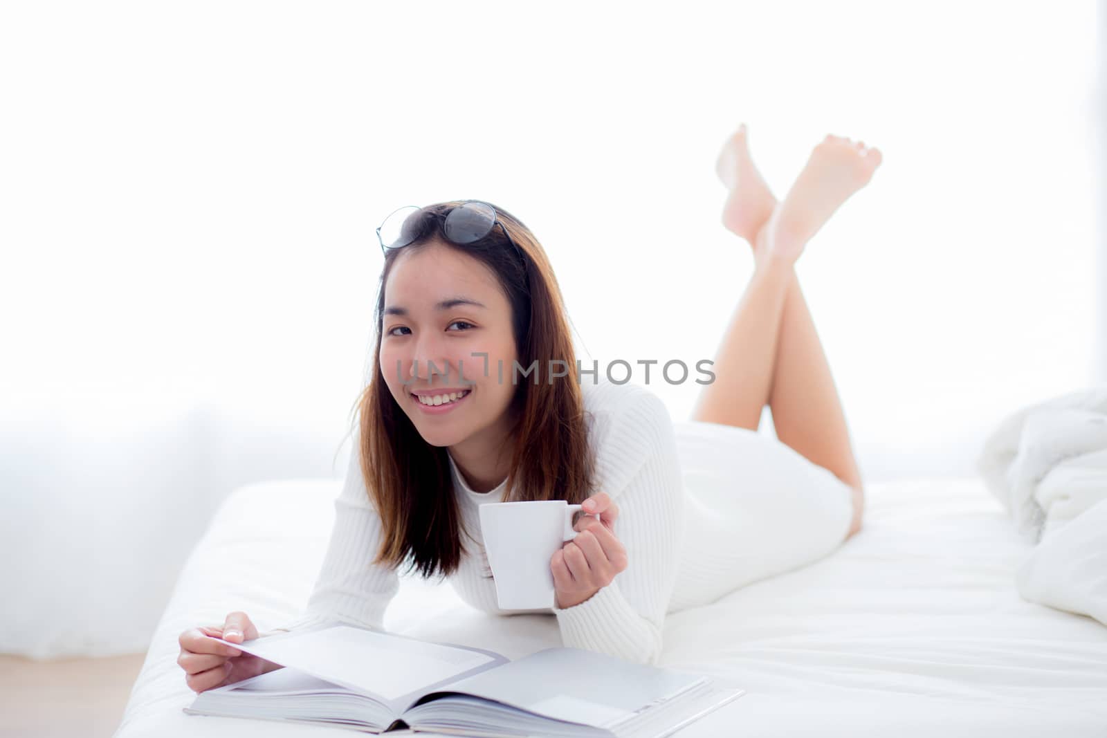 woman lying on bed reading a book and holding coffee cup looking into camera in bedroom.