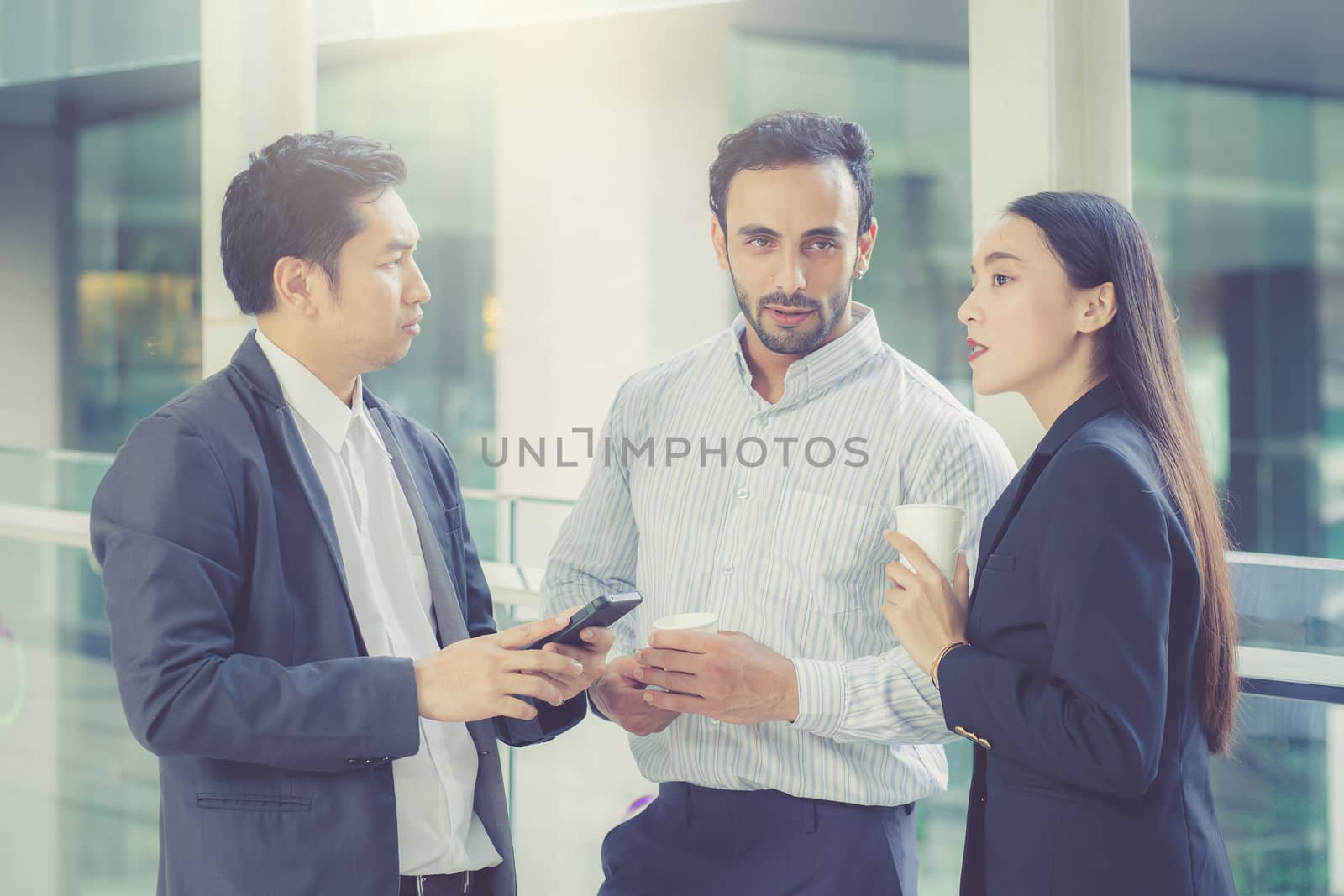 Two handsome young businessmen and lady in classic suits are holding cups of coffee, talking and smiling, standing outside the office building.