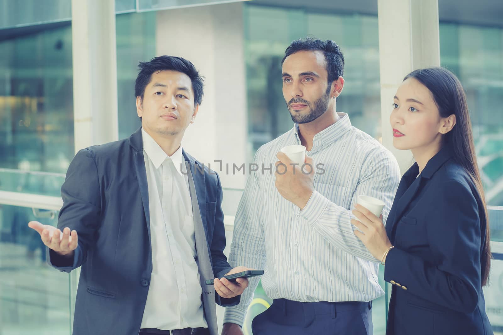 Two handsome young businessmen and lady in classic suits are holding cups of coffee, talking and smiling, standing outside the office building.
