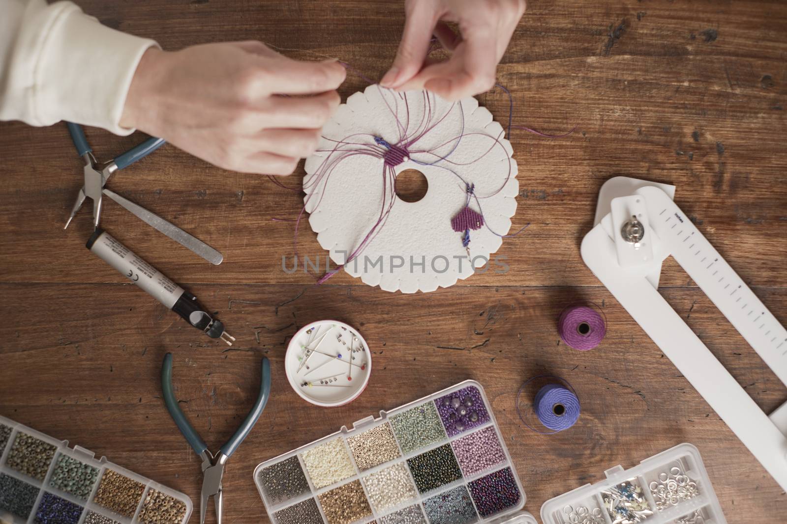 Lifestyle concept, work from home to reinvent your life: top view of woman hands making macrame knots the fuchsia thread creating an earring on kumihimo tools on wooden table by robbyfontanesi