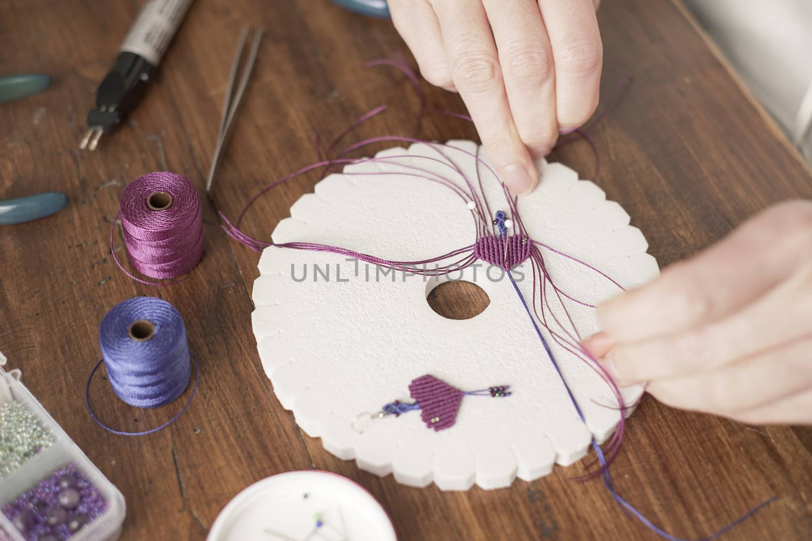 Lifestyle concept, work from home to reinvent your life: close-up of woman hands making macrame knots the fuchsia thread creating an earring on kumihimo tools on wooden table