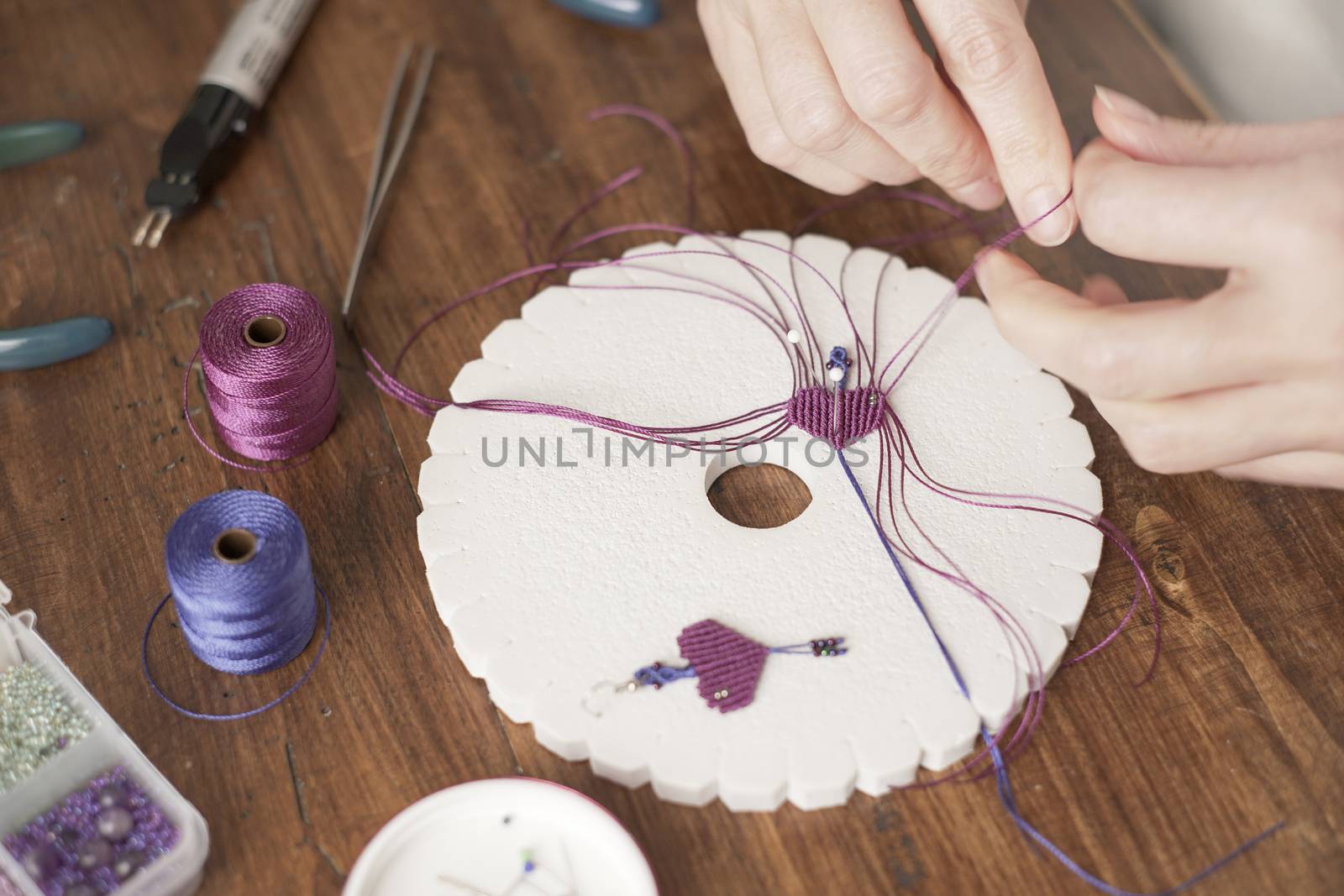 Lifestyle concept, work from home to reinvent your life: close-up of woman hands making macrame knots the fuchsia thread creating an earring on kumihimo tools on wooden table