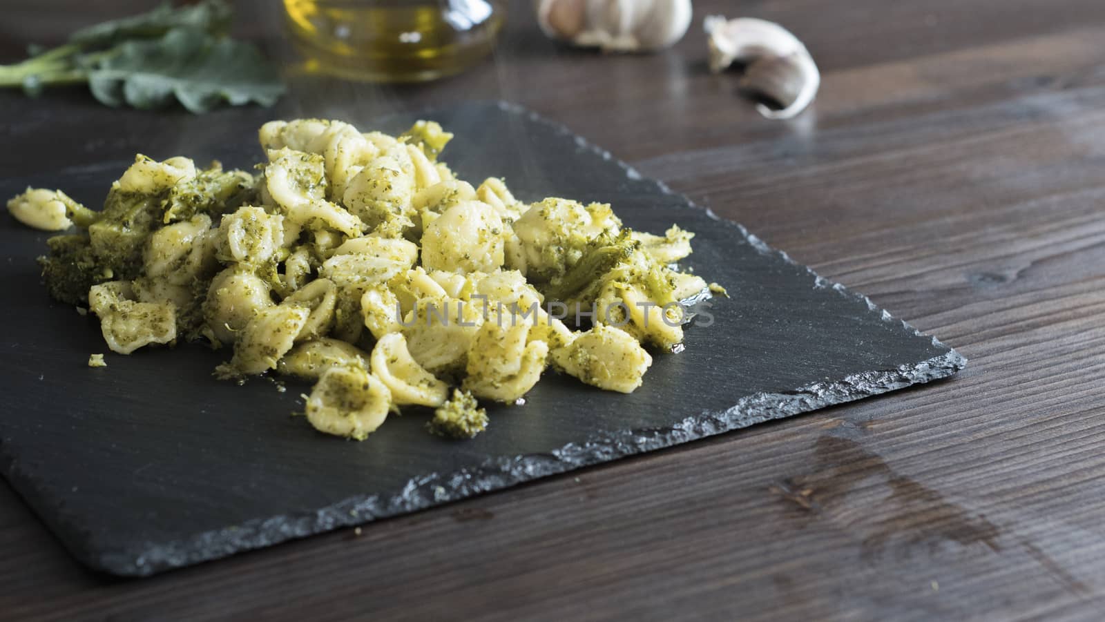 Closeup of a slate plate with steaming orecchiette with broccoli, typical Apulian recipe, in backlight on dark wooden table with olive oil and garlic on background in bokeh effect