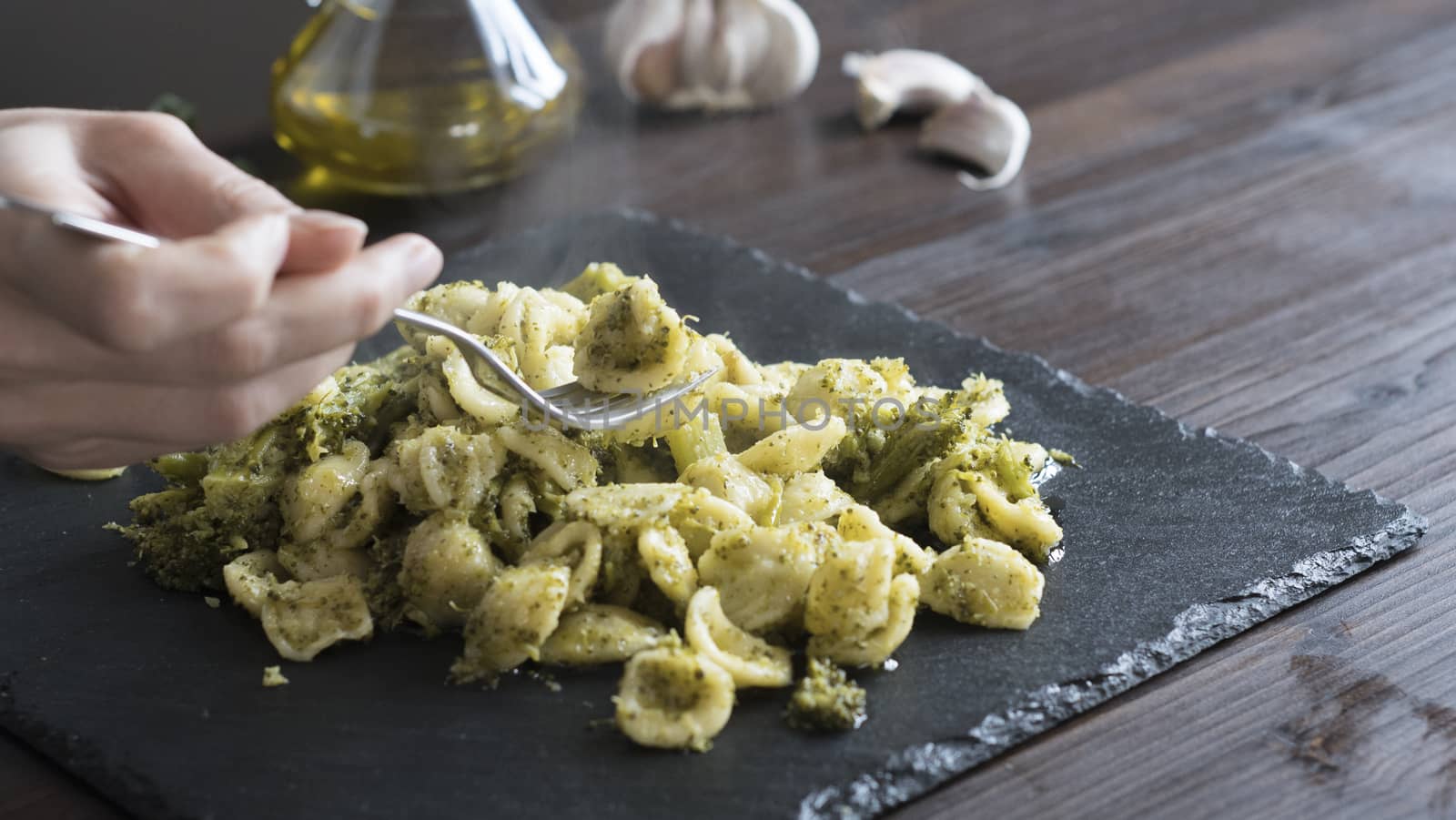 Closeup of a woman's hand that pick up with a fork steaming orecchiette with broccoli, typical Apulian recipe, in backlight on dark wooden table with olive oil and garlic on background in bokeh effect by robbyfontanesi