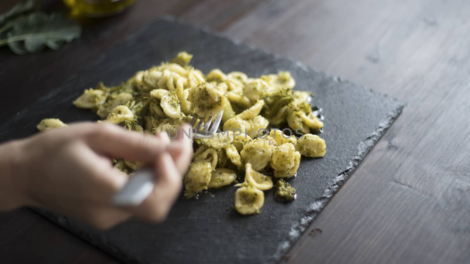 Closeup of a woman's hand that pick up with a fork steaming orecchiette with broccoli, typical Apulian recipe, in backlight on dark wooden table with olive oil and garlic on background in bokeh effect