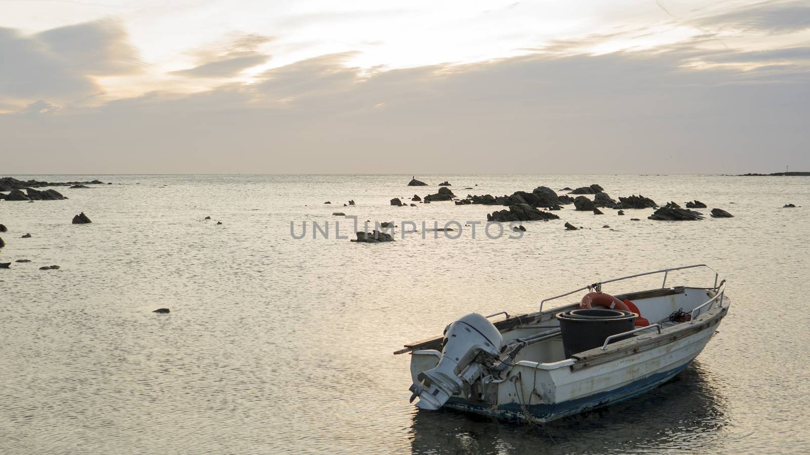 Emotional shot of the sea at dawn in a small beach with cloudy sky and a fishing boat anchored to the shore with a rope moving in flat water by robbyfontanesi