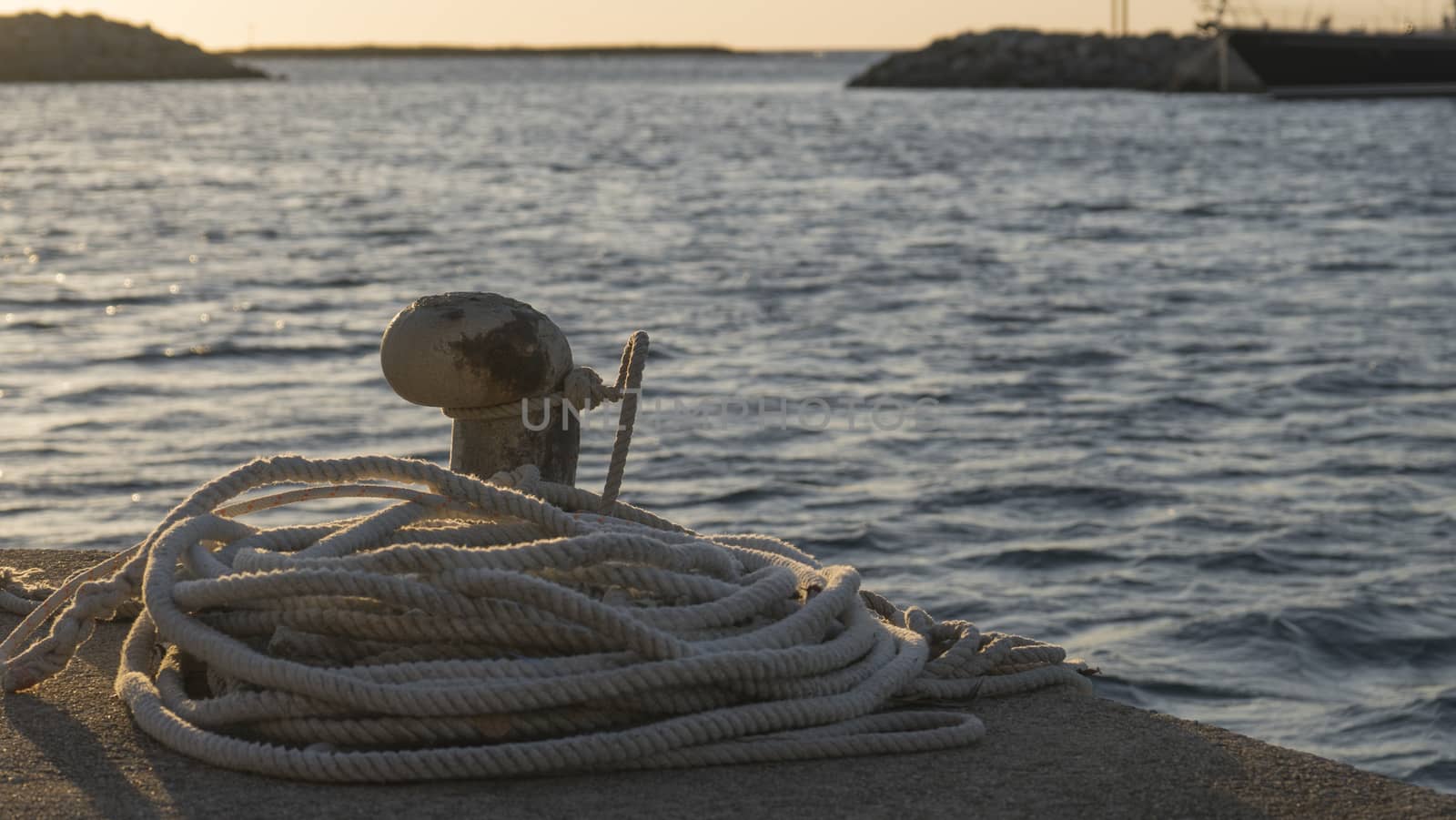 Backlight foreground at sunrise or sunset of a mooring bollard with the rope tied and resting on the ground and various boats in the background in back light by robbyfontanesi