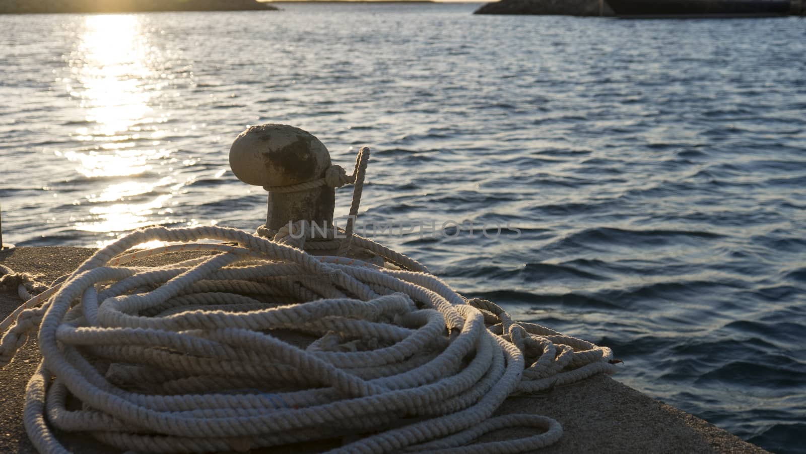 Backlight foreground at sunrise or sunset of a mooring bollard with the rope tied and resting on the ground and various boats in the background in back light by robbyfontanesi