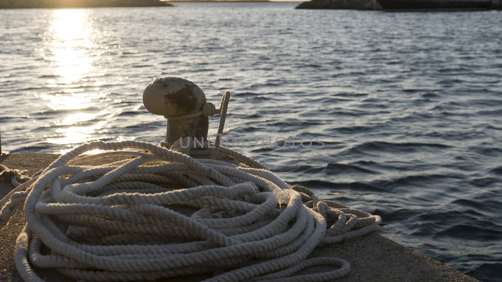 Backlight foreground at sunrise or sunset of a mooring bollard with the rope tied and resting on the ground and various boats in the background in back light by robbyfontanesi