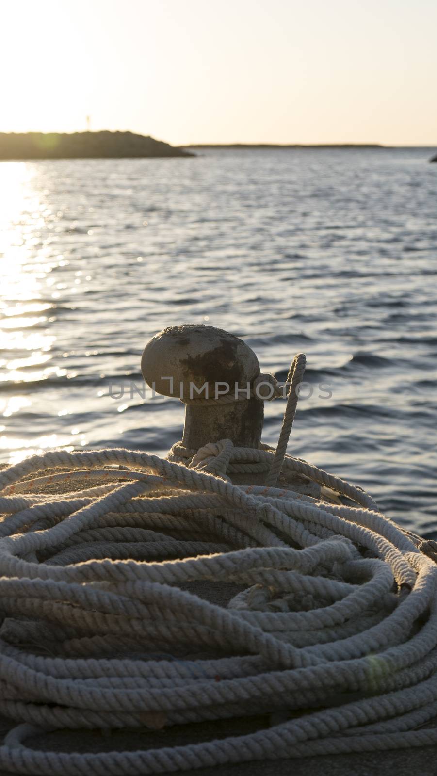 Backlight foreground at sunrise or sunset of a mooring bollard with the rope tied and resting on the ground and various boats in the background in back light by robbyfontanesi