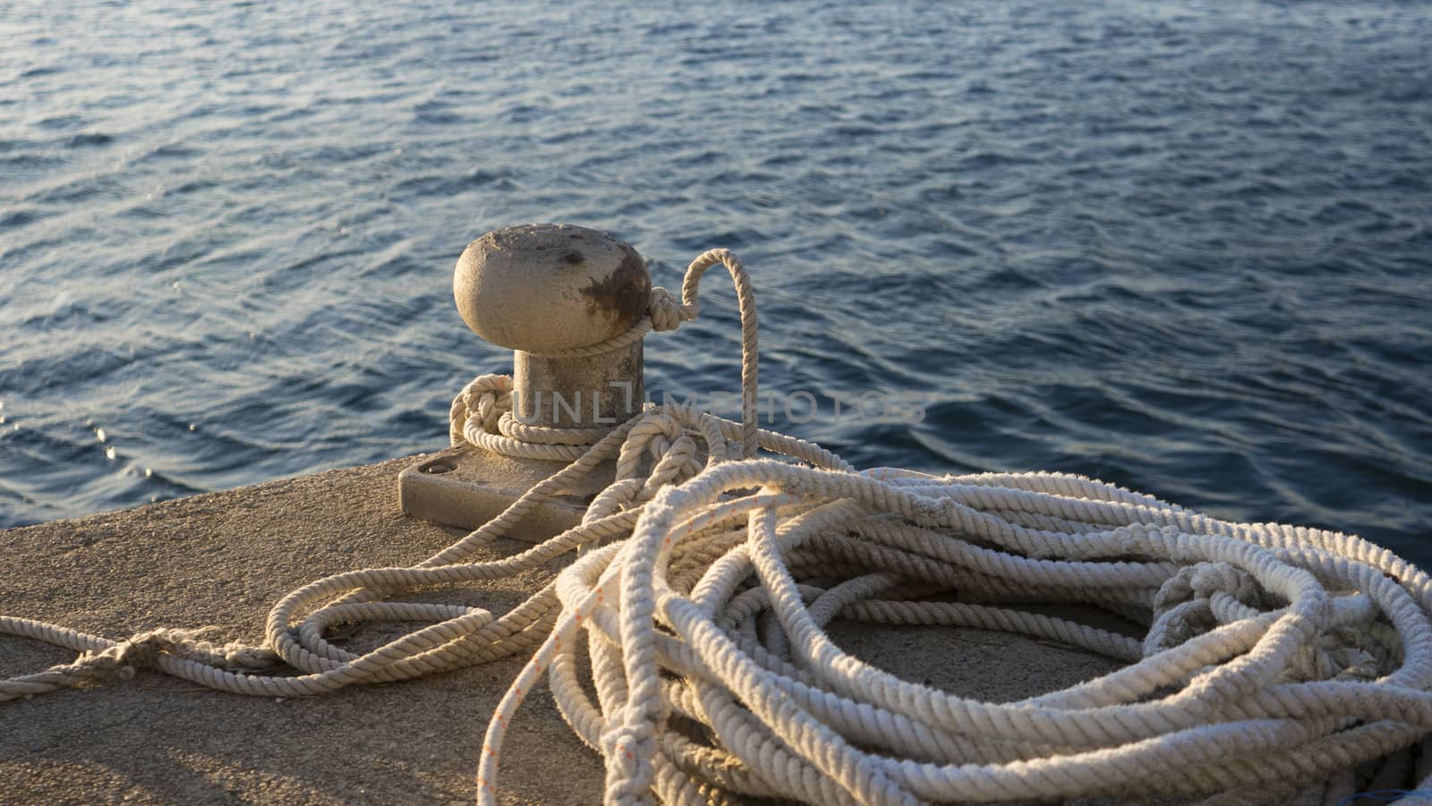 Backlight foreground at sunrise or sunset of a mooring bollard with the rope tied and resting on the ground and various boats in the background in back light