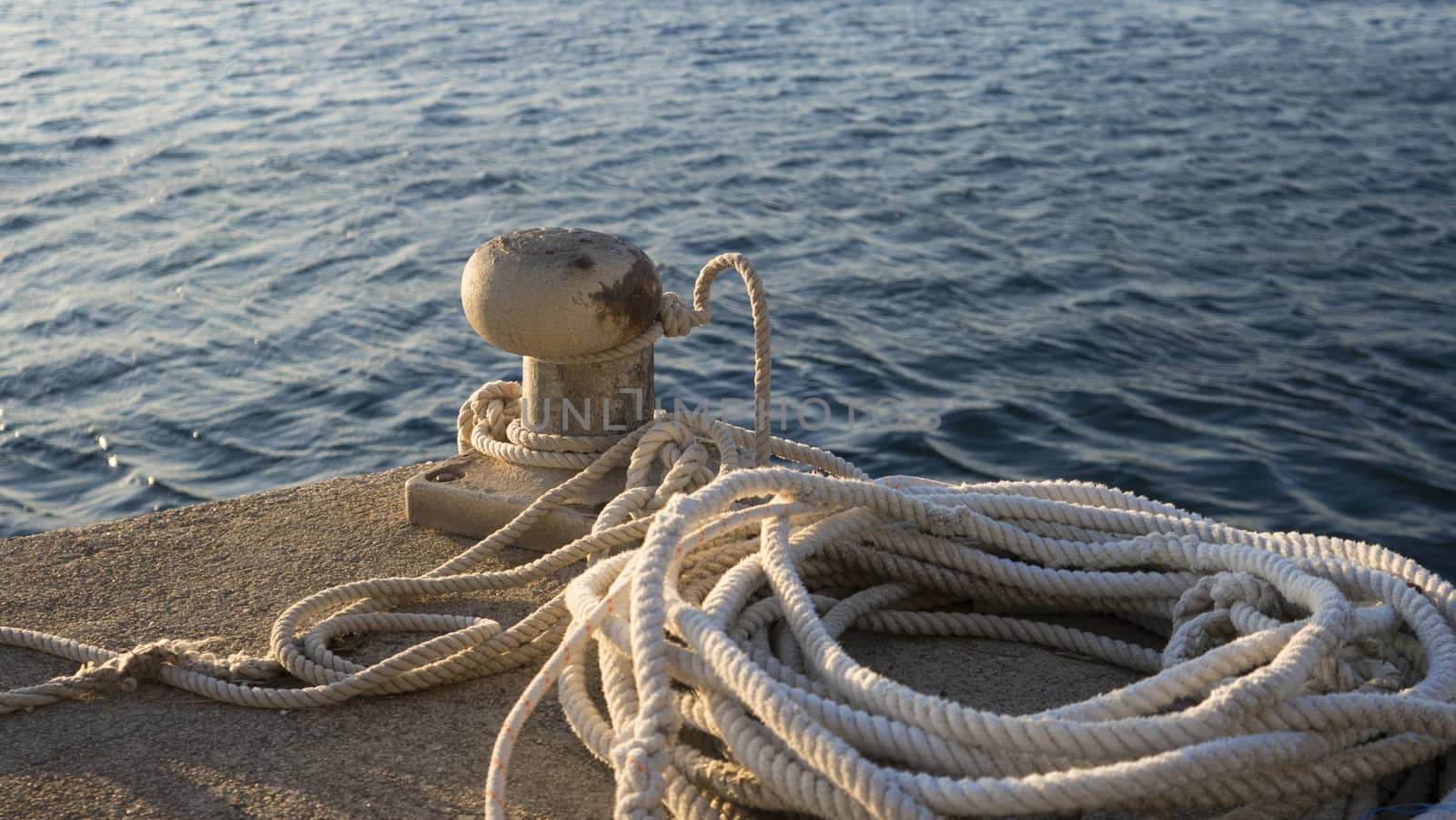 Backlight foreground at sunrise or sunset of a mooring bollard with the rope tied and resting on the ground and various boats in the background in back light by robbyfontanesi