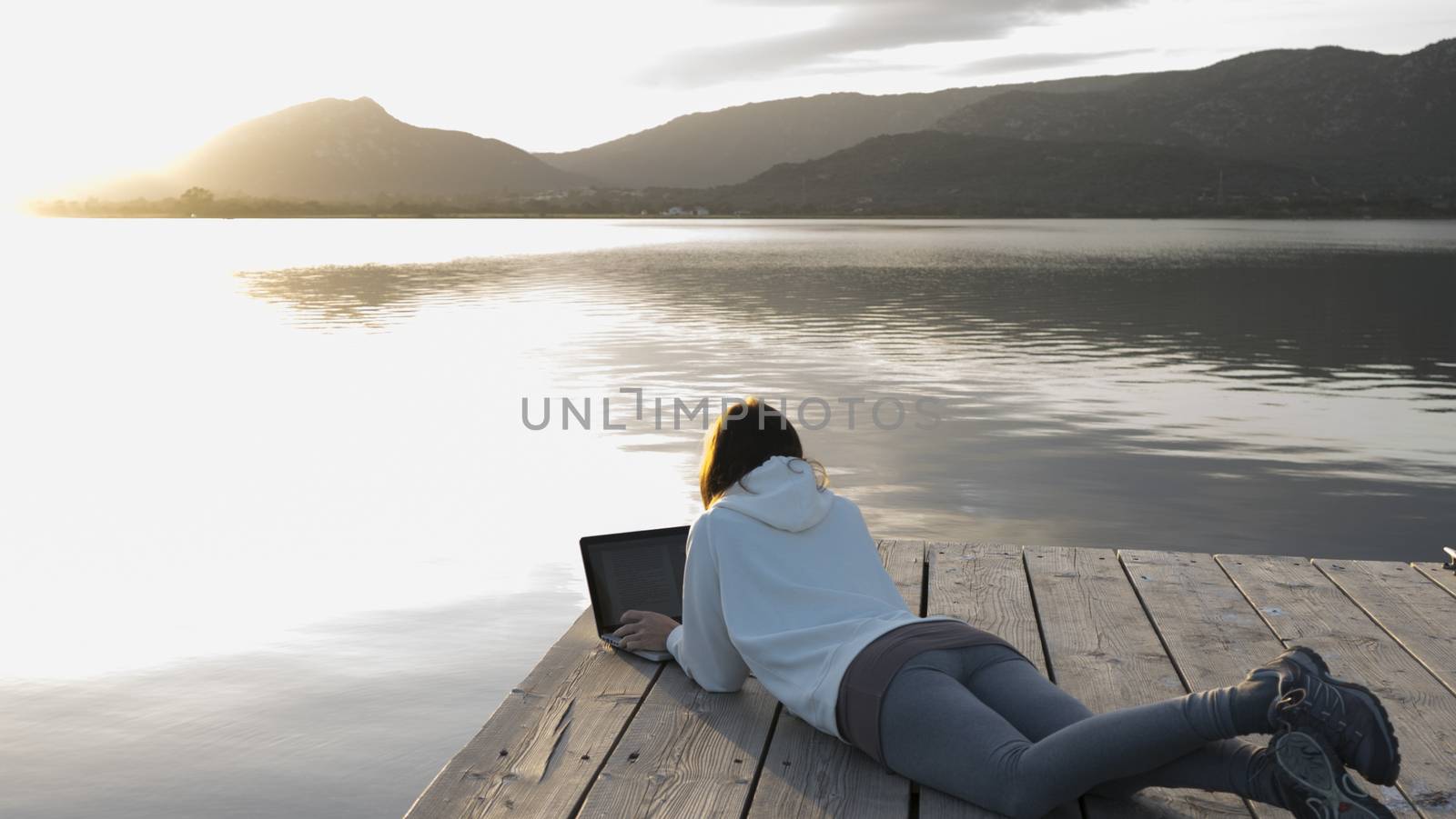 Internet freelance job choice concept: a young woman works on her laptop lying on a pier by a lake at sunset