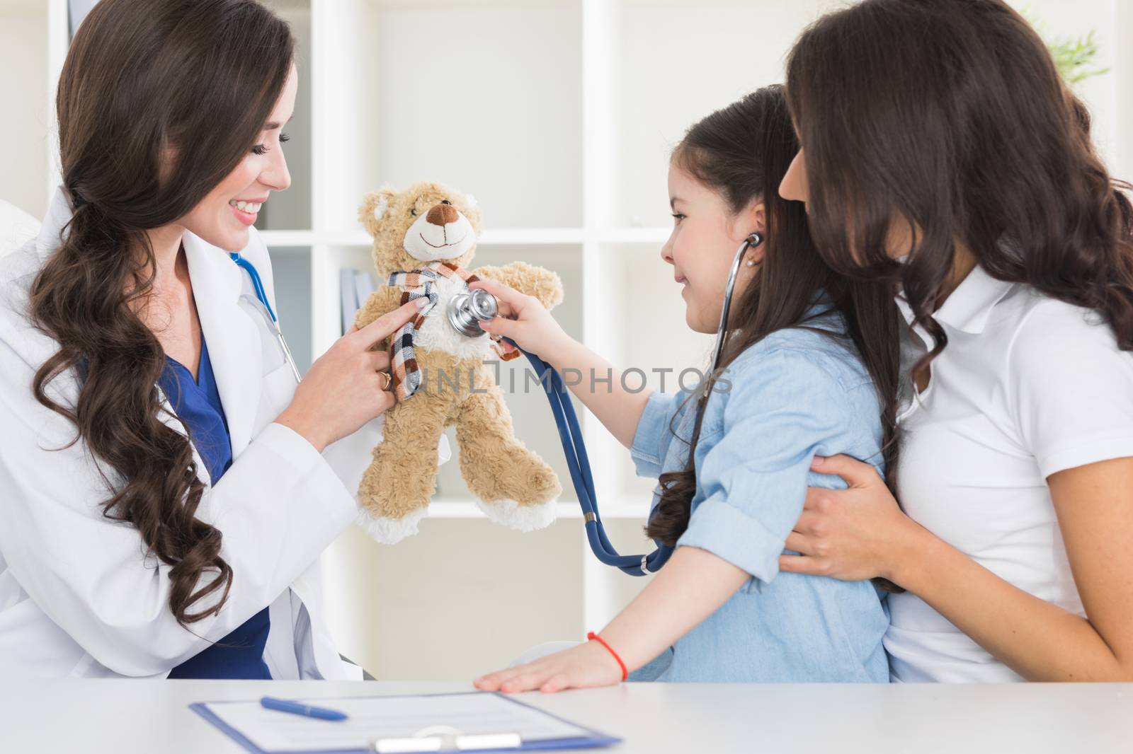 Cute small child patient pretending to be doctor, holding teddy bear toy at meeting with general practitioner. Friendly nurse showing how stethoscope works to smiling little girl at checkup in clinic.