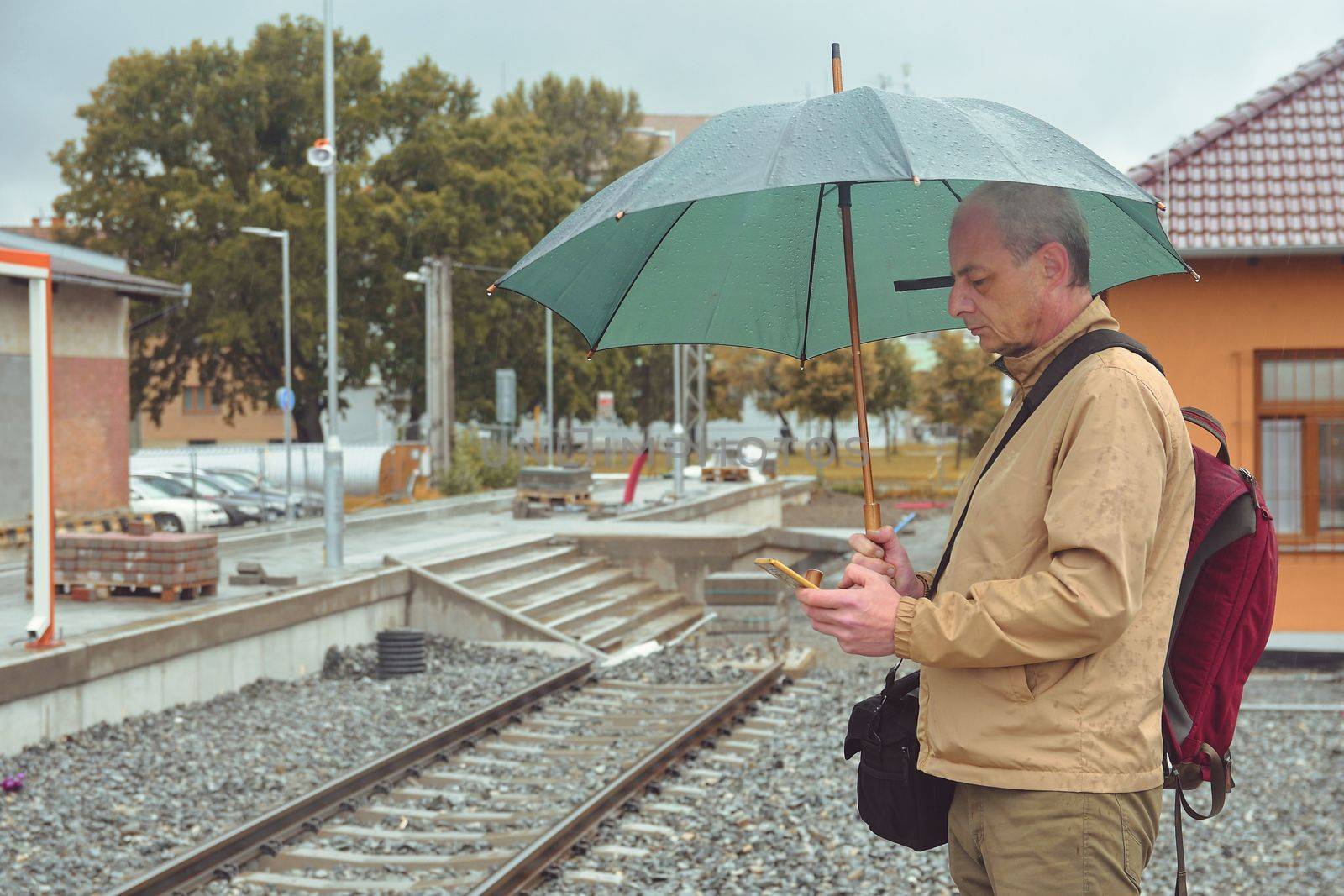 Portrait of a mature traveller texting on smartphone with an umbrella standing on the platform of a train station. Unfinished railway line. When the train arrives.. Funny concept of futile situation by roman_nerud