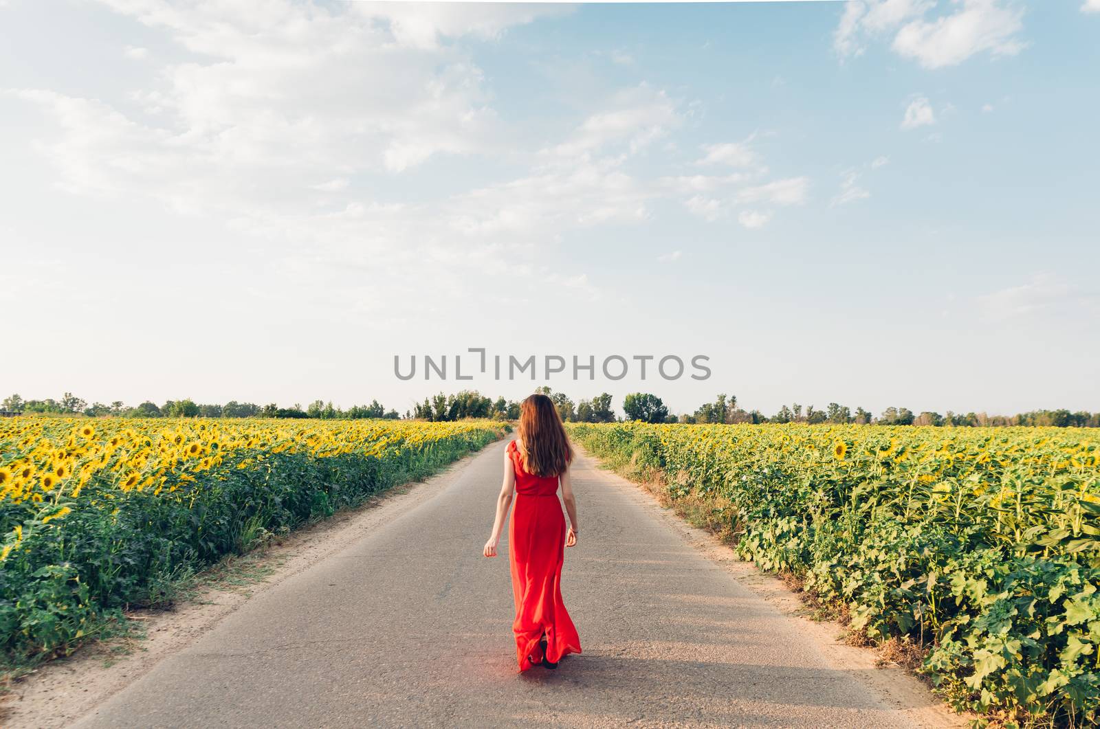 woman with red dress walking on road with sunflowers. by Fotoeventis