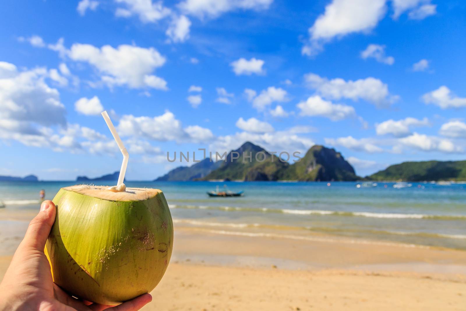 Hand holding a coconut coco loco coctail with straw, tropical islands, beach, sea and blue sky in the background, El Nido, Palawan, Phillipines