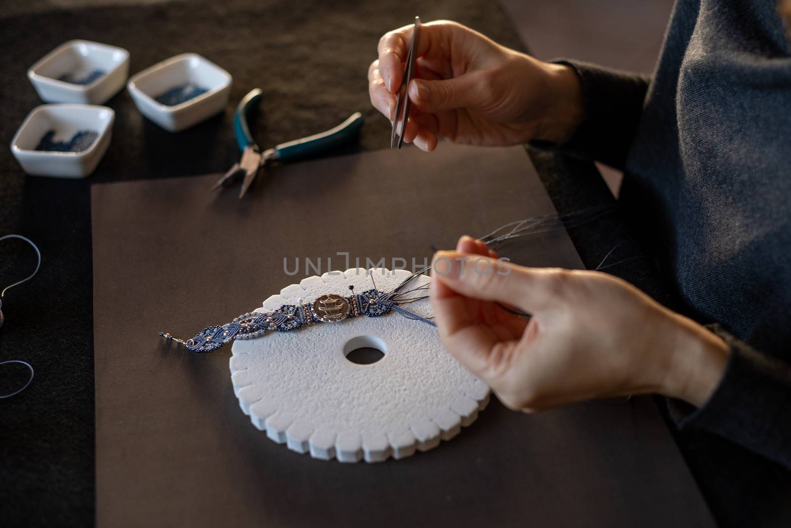 Lifestyle concept, reinvent your life and your job: close-up detail of woman hands making macrame knotted jewel with the fingers threading the bead into the nylon thread on black work table