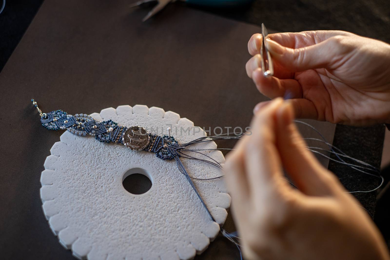 Lifestyle concept, reinvent your life and your job: close-up detail of woman hands making macrame knotted jewel with the fingers threading the bead into the nylon thread on black work table by robbyfontanesi