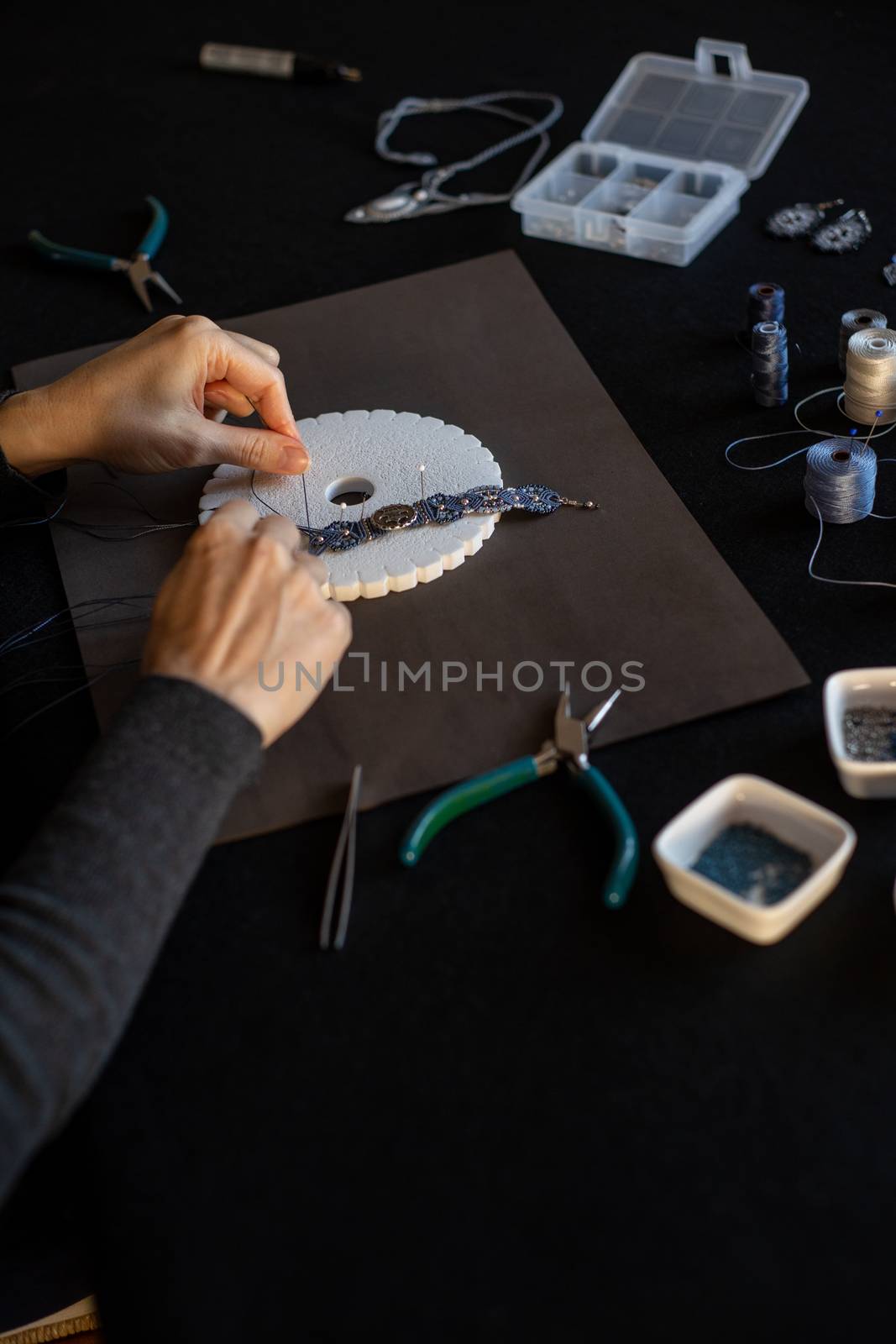 Lifestyle concept, work from home to reinvent your life: close-up of woman hands making macrame knotted jewelry with stone beads and tools on black work table by robbyfontanesi