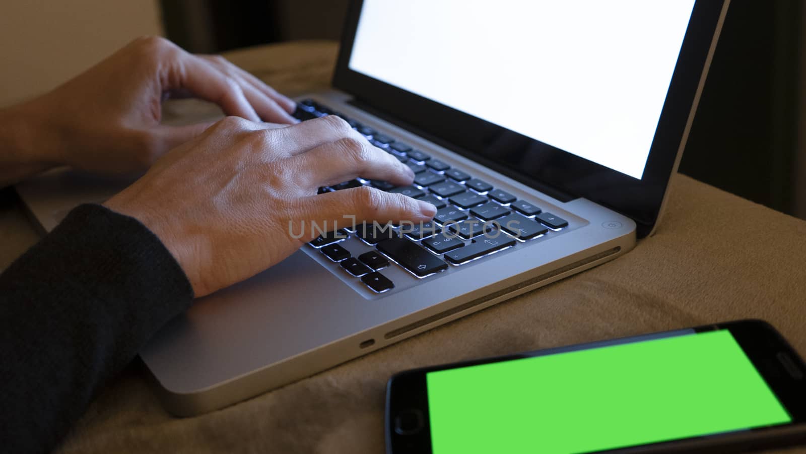 Couple relationships and dating in modern times concept: closeup of woman's hands typing on laptop keyboard with blank white screen in low light with smartphone alongside with green blank screen by robbyfontanesi