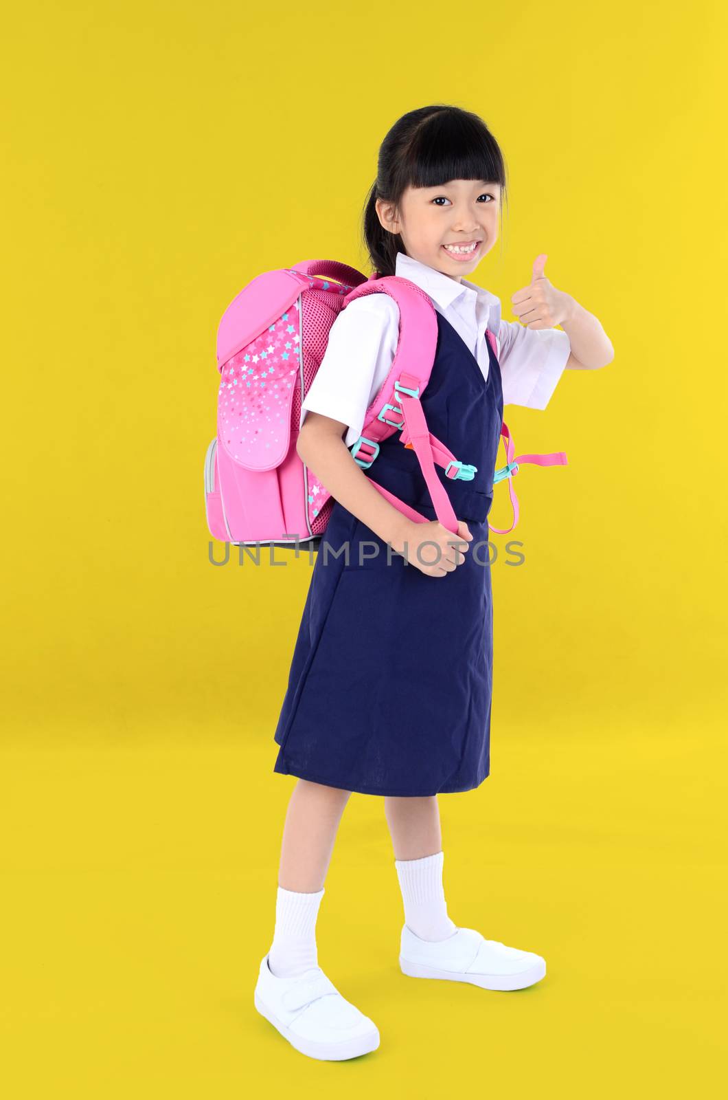 Back to school. Portrait of happy asian cute little child girl in uniform with school bag smiling  show finger thumb up when go back to school, isolated orange background. 