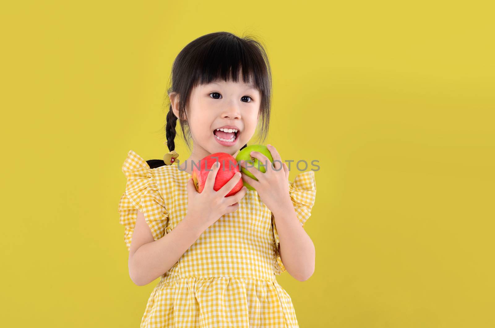 happy little asian girl holding green and red apple isolated on yellow background.