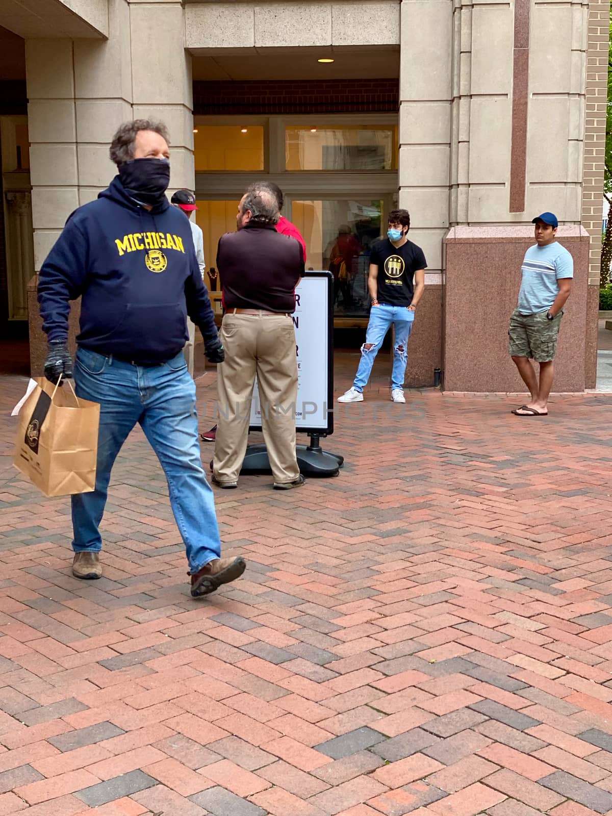 Reston, VA, USA -- April 25, 2020. A man wearing a face mask carries his take-out dinner in a bag; others wait for thier orders to be fulfilled.