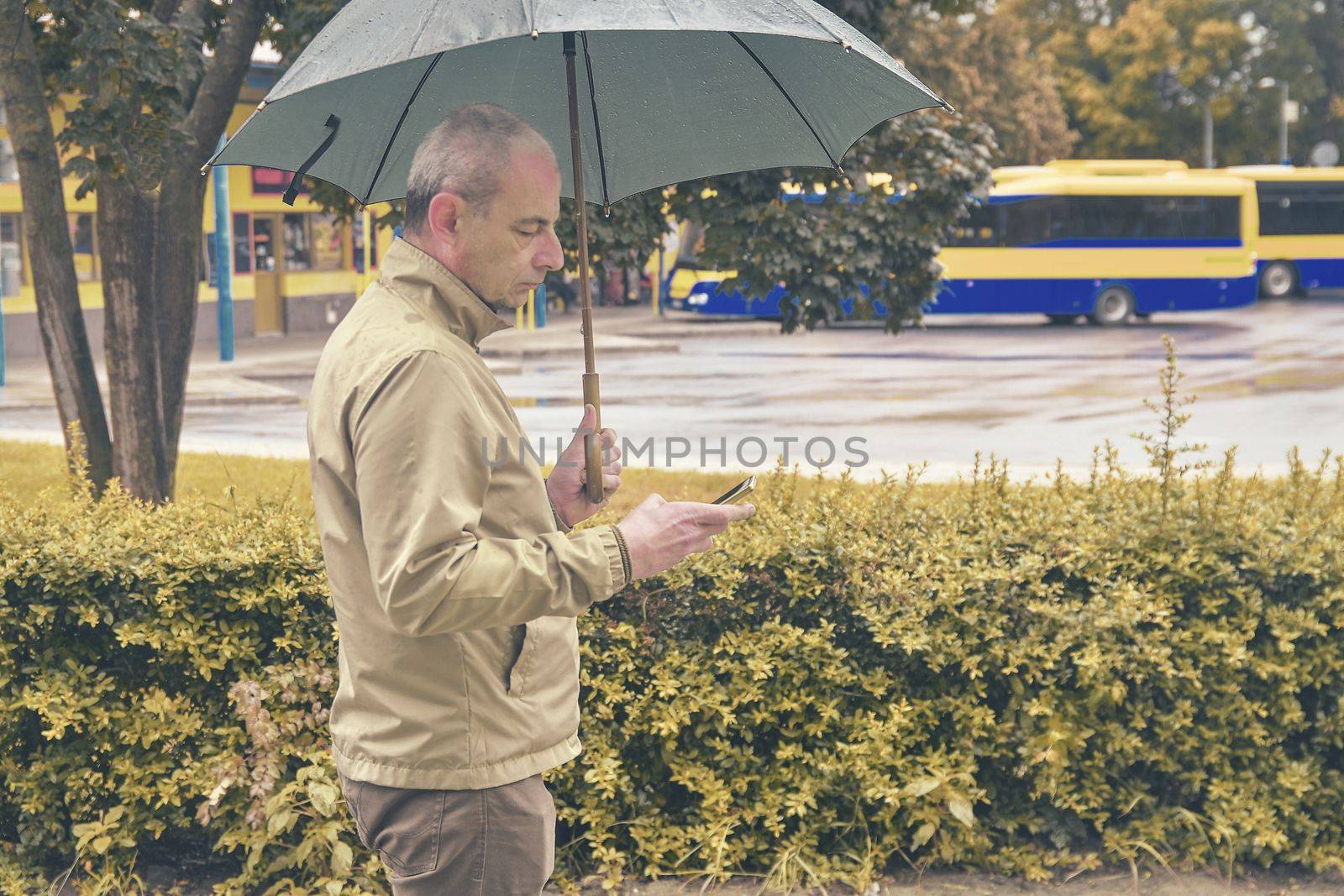 A man standing at the bus station with an umbrella waiting for a bus. Man waiting on a bus, texting on mobile phone in a rainy day. Rainy day in a local town.