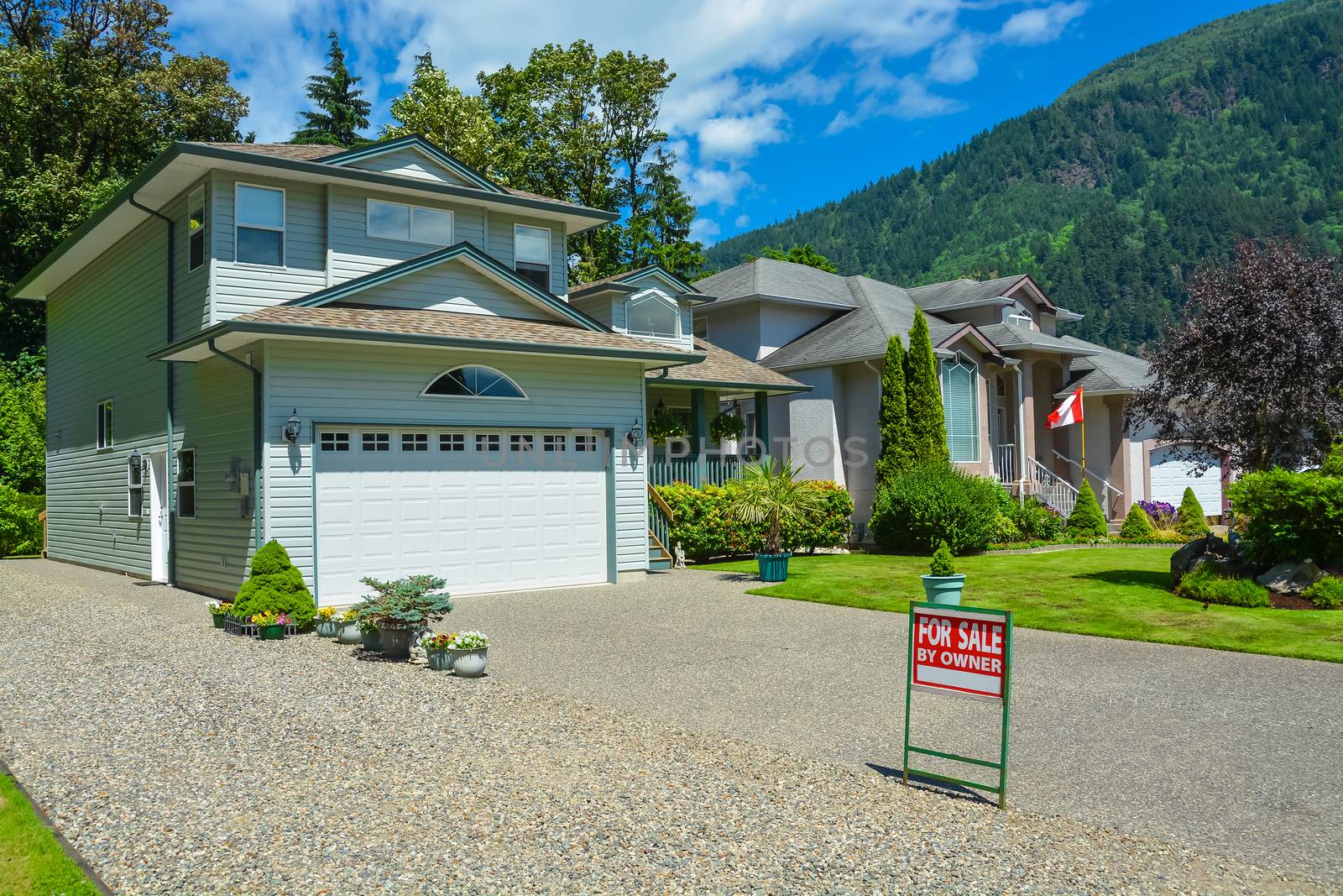 Suburban house in British Columbia for sale. Big house with wide garage door, concrete driveway, and lawn in front for sale. House with mountain view and blue sky background.