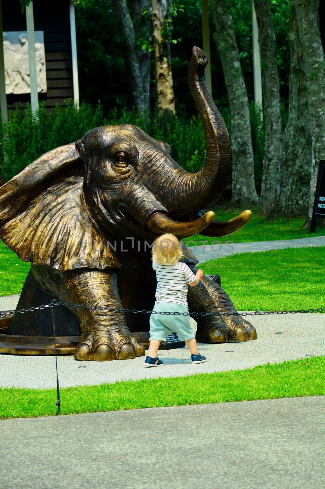 Matakana, New Zealand - Dec 2019: Sculptureum sculpture park. Bronze sculpture representing an elephant climbing out of a manhole, and a cute little child fascinated by the elephant. by Marshalkina