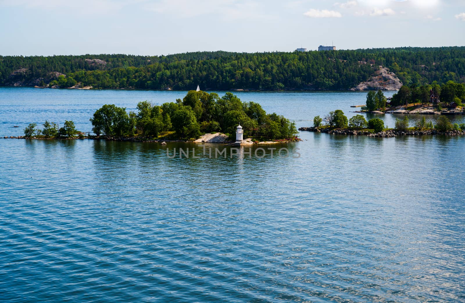 A white lighthouse surrounded by green trees on a small island in the deep blue sea of the Swedish archipelago.