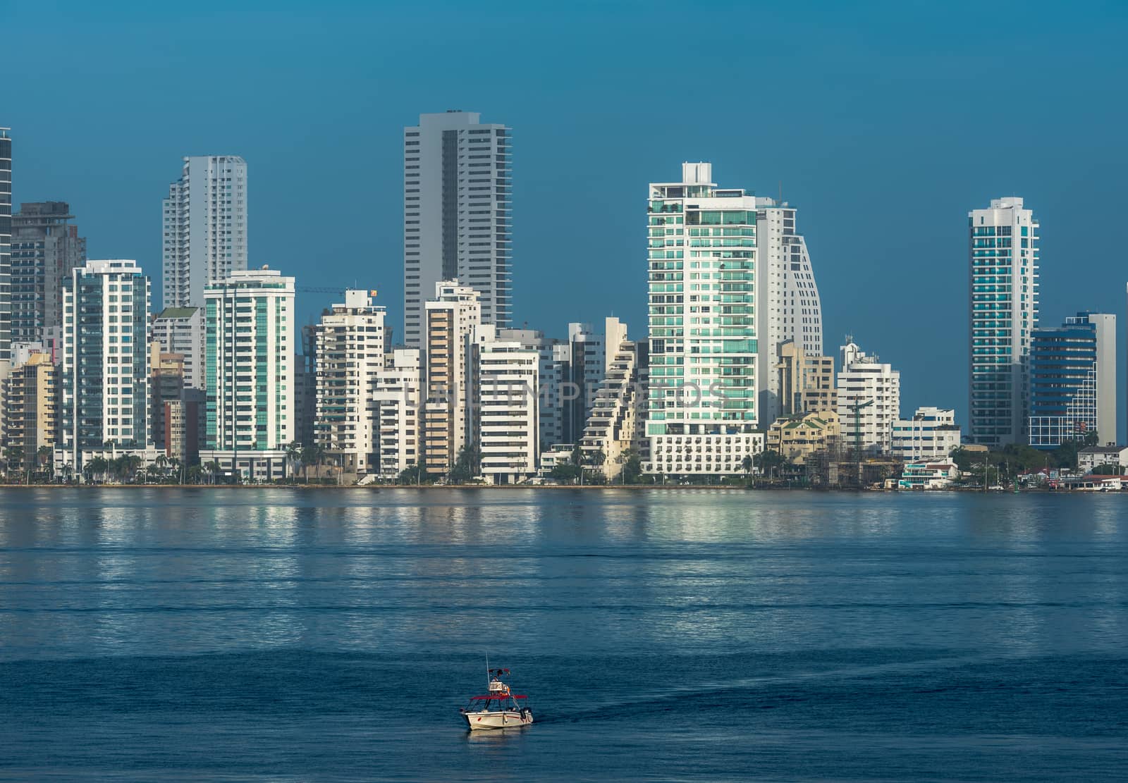 A small boat sails on the blue waters of Cartagena Bay with the city's skyscrapers gleaming in the background.