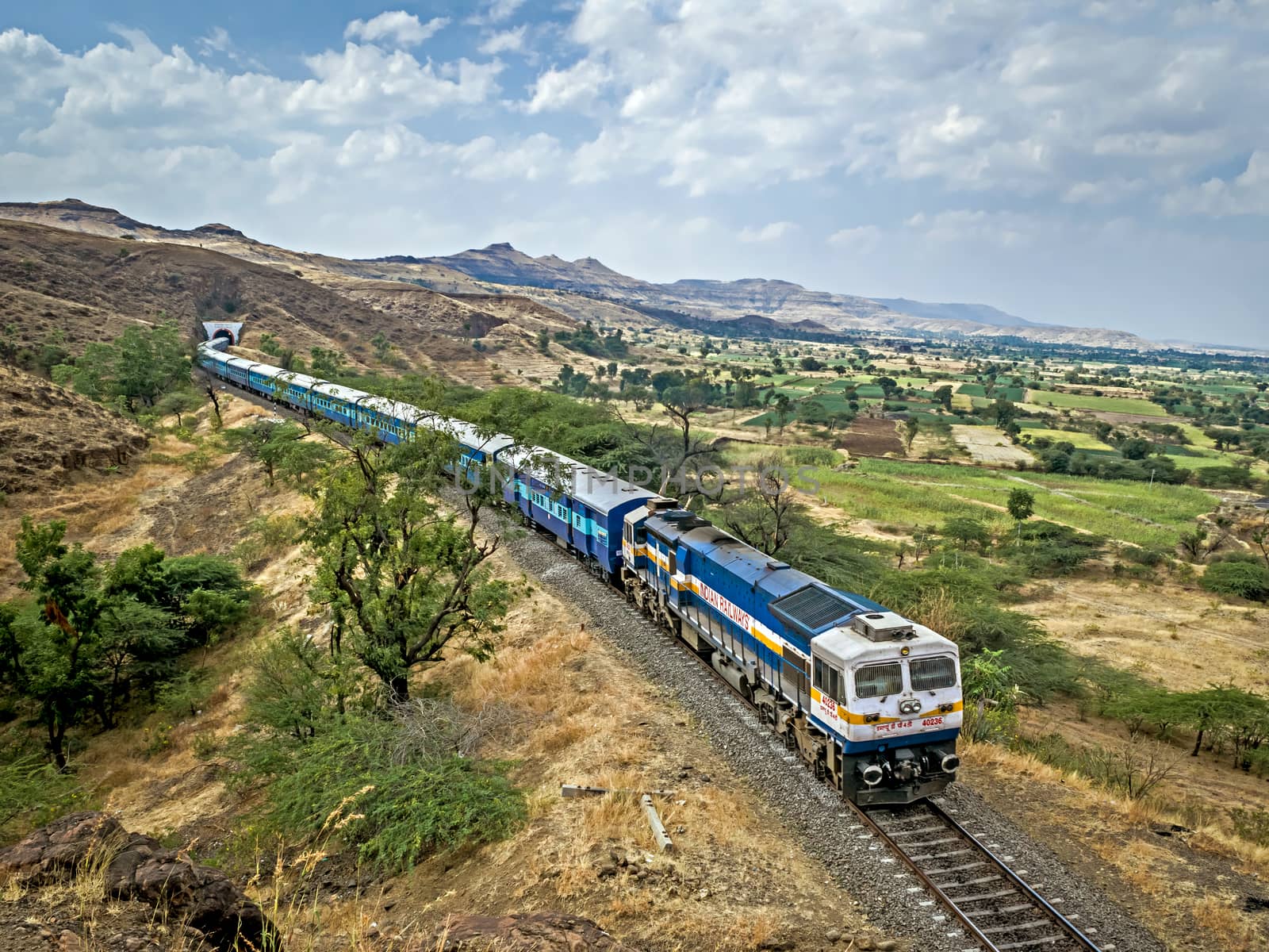 Shindawane, Pune, India:January 16th, 2016-A long train exits a tunnel on a clear background of blue sky with clouds. by lalam