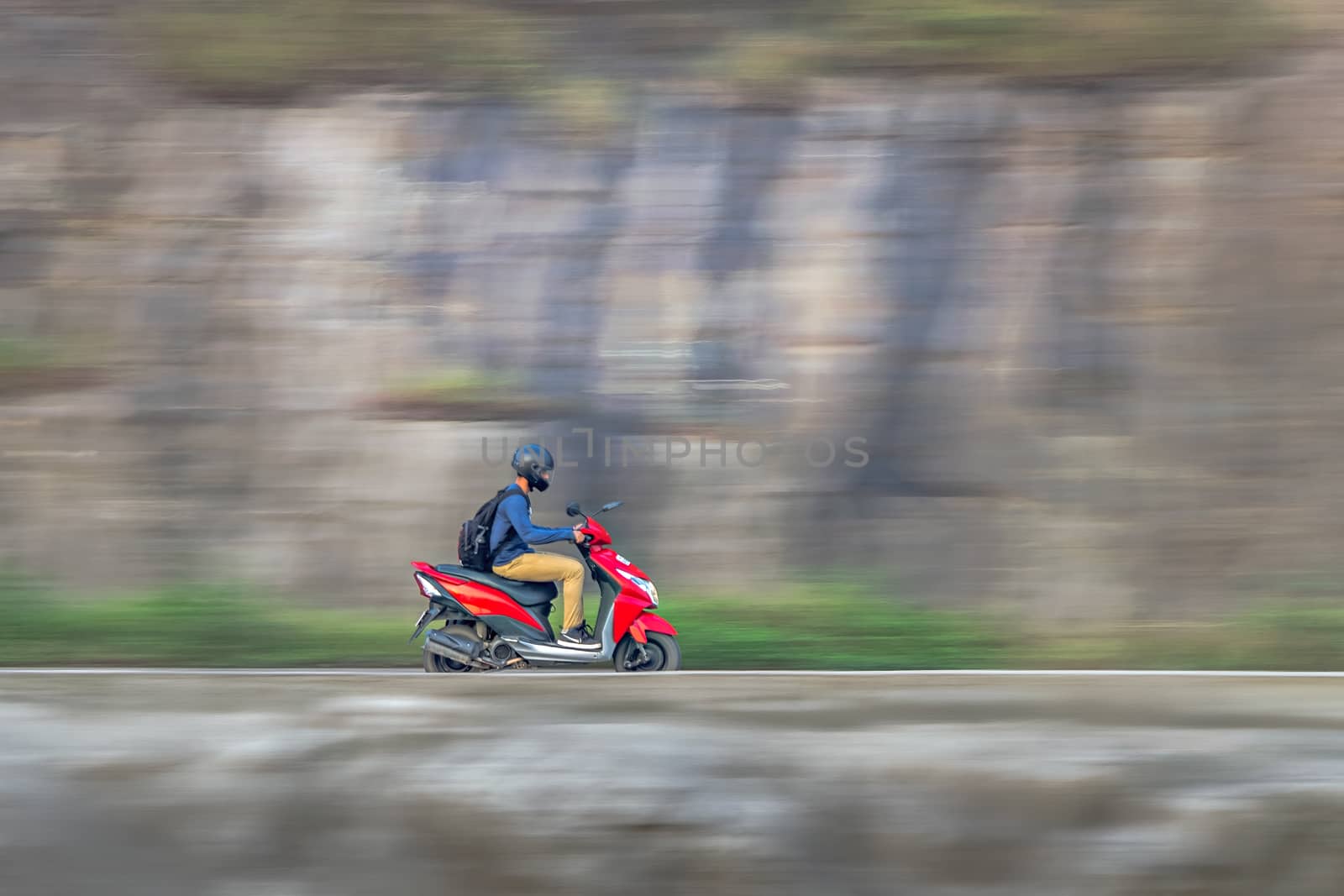Motion blur image of a rider wearing helmet for safety, riding uphill on a red two wheeler moped.