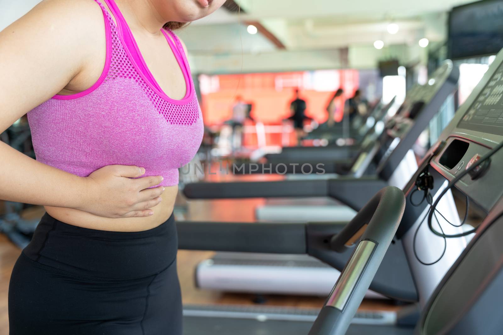 A woman feeling exhausted and suffering from stomach side pain and injury while running on treadmill at fitness gym. Sport, health care and medical concept.