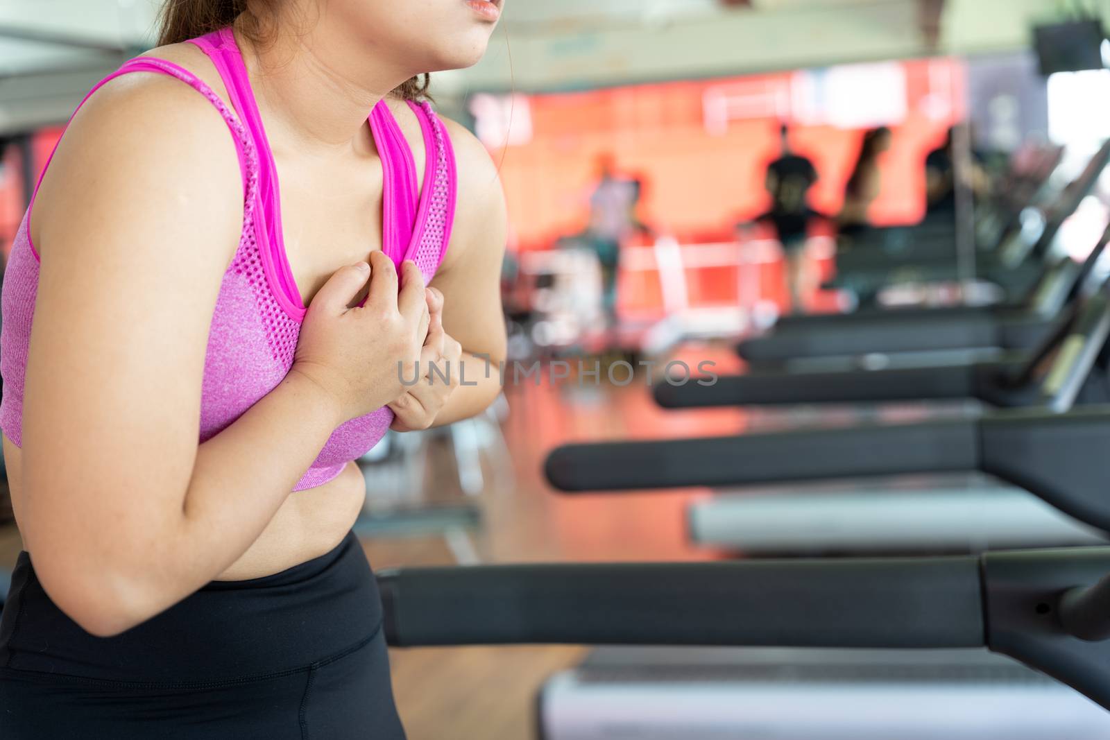 A woman feeling exhausted and suffering from heart pain and injury while running on treadmill at fitness gym. Sport, health care and medical concept.