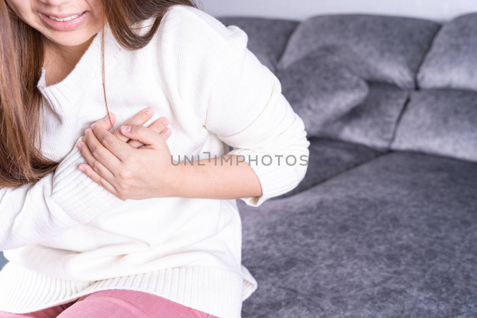 Young woman suffering from heart pain while sitting on sofa at home. Healthcare medical or daily life concept.