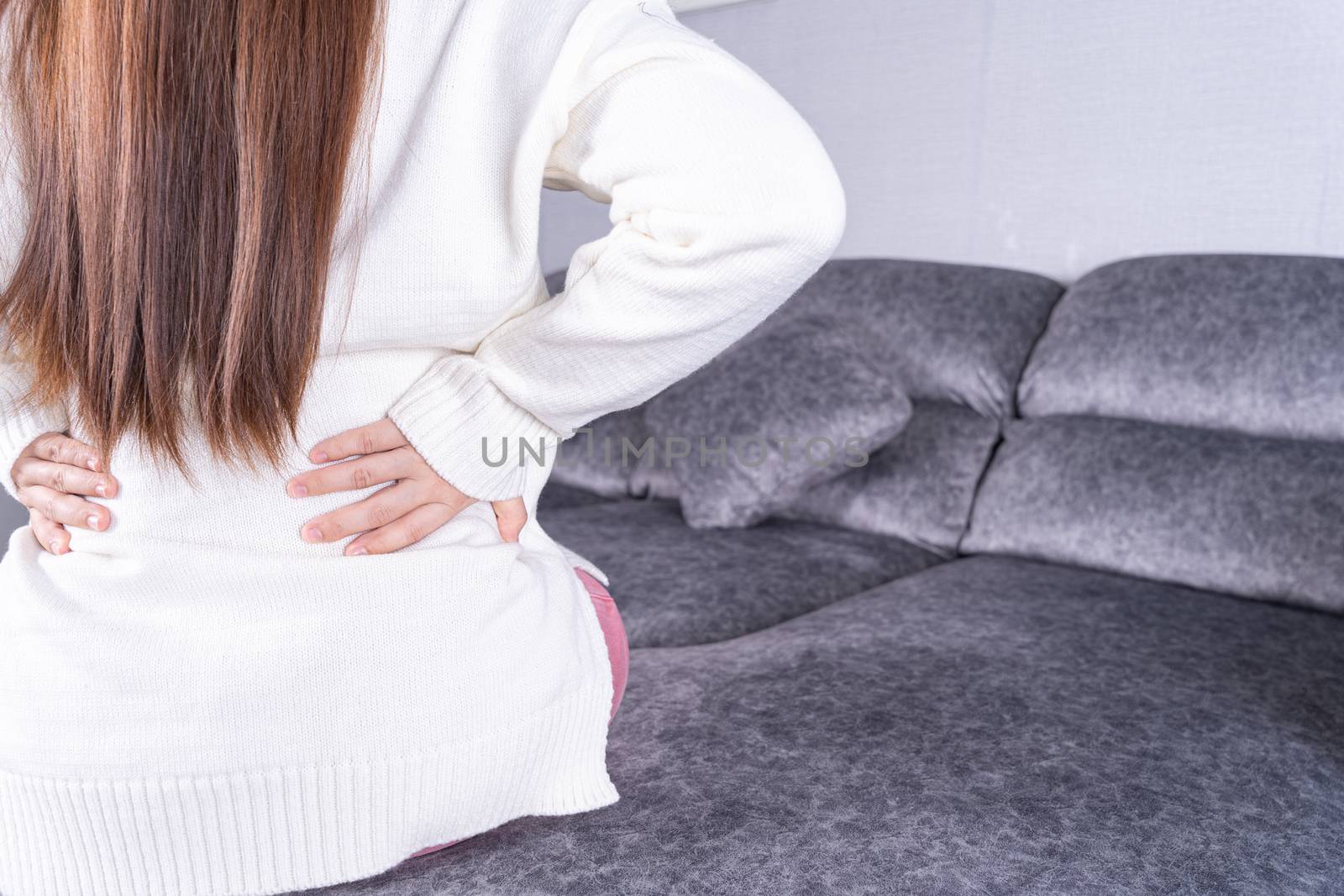 Young woman suffering from back pain while sitting on sofa at home. Healthcare medical or daily life concept.