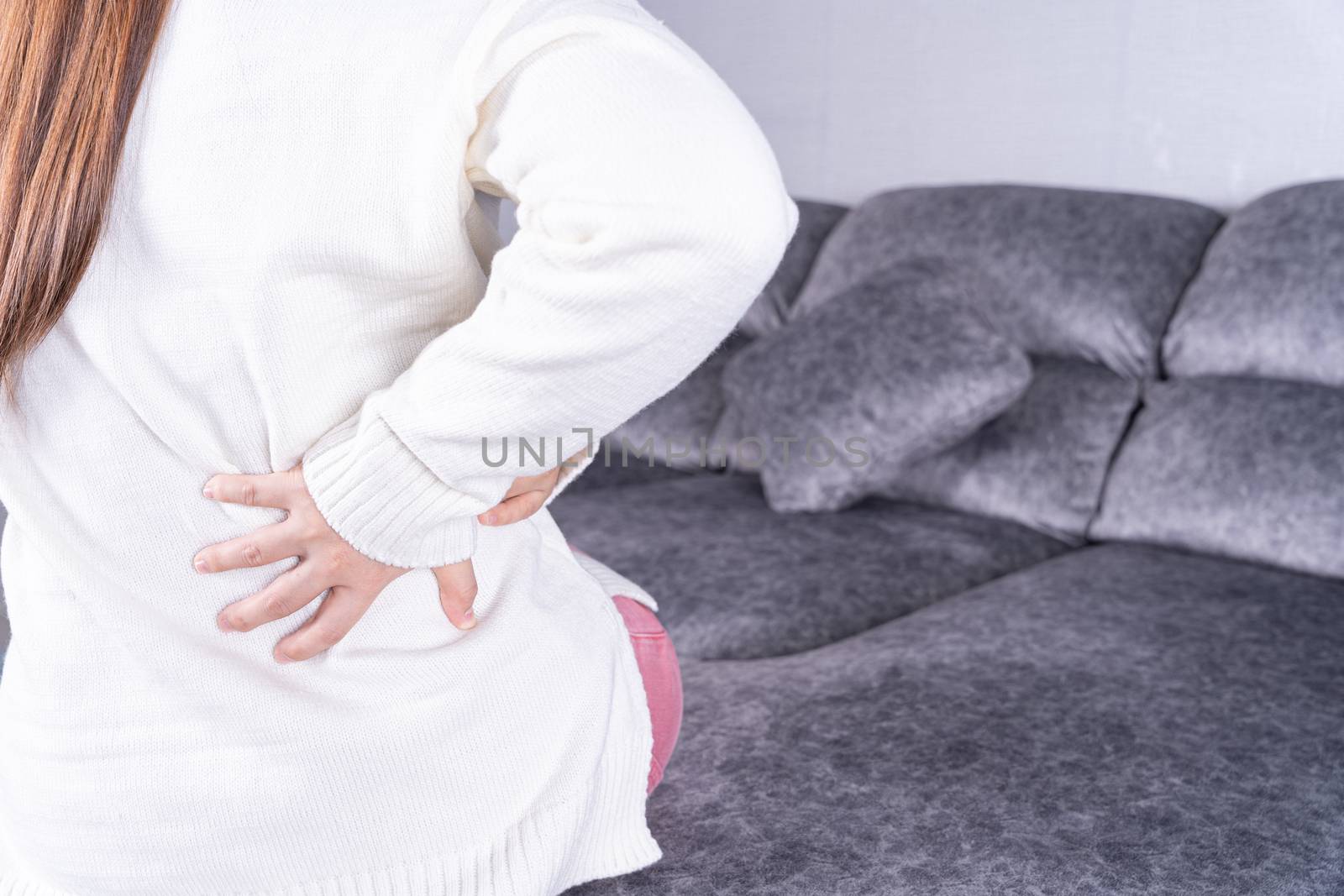 Young woman suffering from back pain while sitting on sofa at home. Healthcare medical or daily life concept.