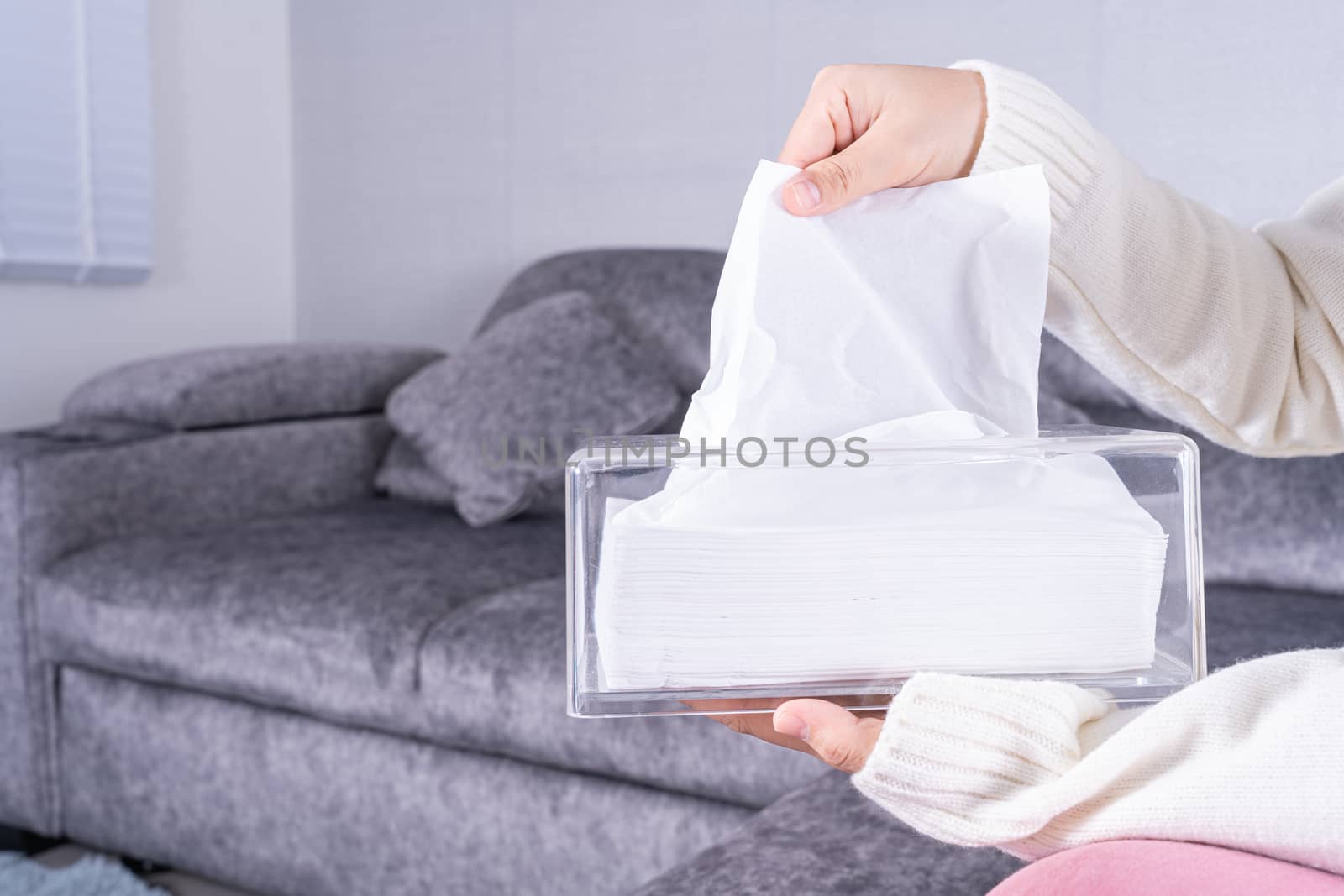 Delicate female hands pulling a white tissue paper out of a transparent crystal tissue box on sofa at home.