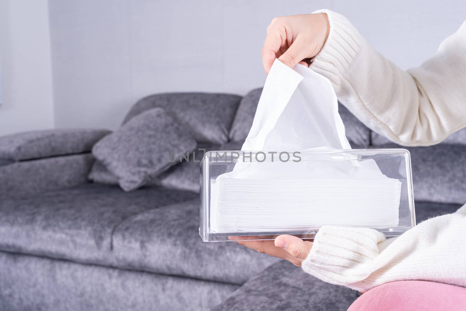 Delicate female hands pulling a white tissue paper out of a transparent crystal tissue box on sofa at home.